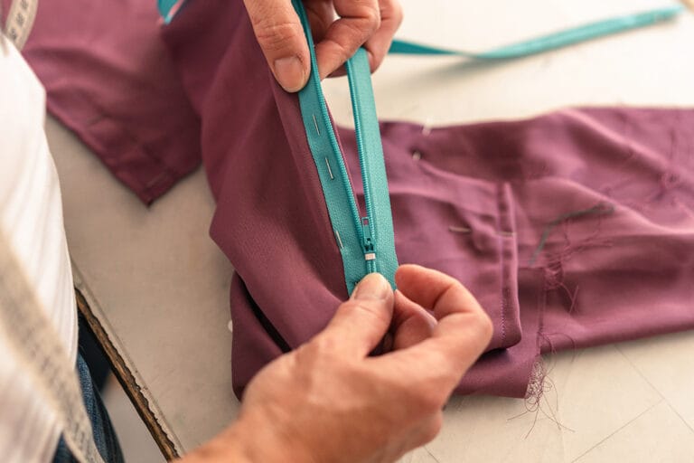 Person attaching a turquoise zipper to purple fabric – hands sewing zipper with pins visible.