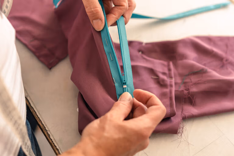 Person attaching a turquoise zipper to purple fabric – hands sewing zipper with pins visible.