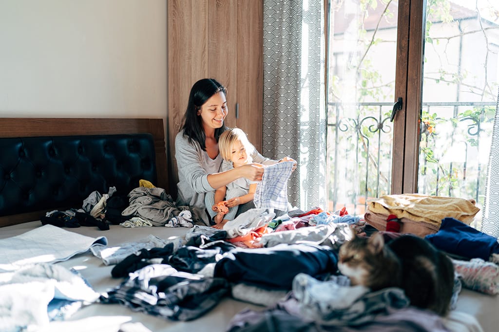 Woman and child folding clothes on bed – bright natural light coming through window.