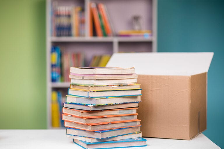 A tall stack of colourful books next to an open cardboard box, with bookshelves in the background.