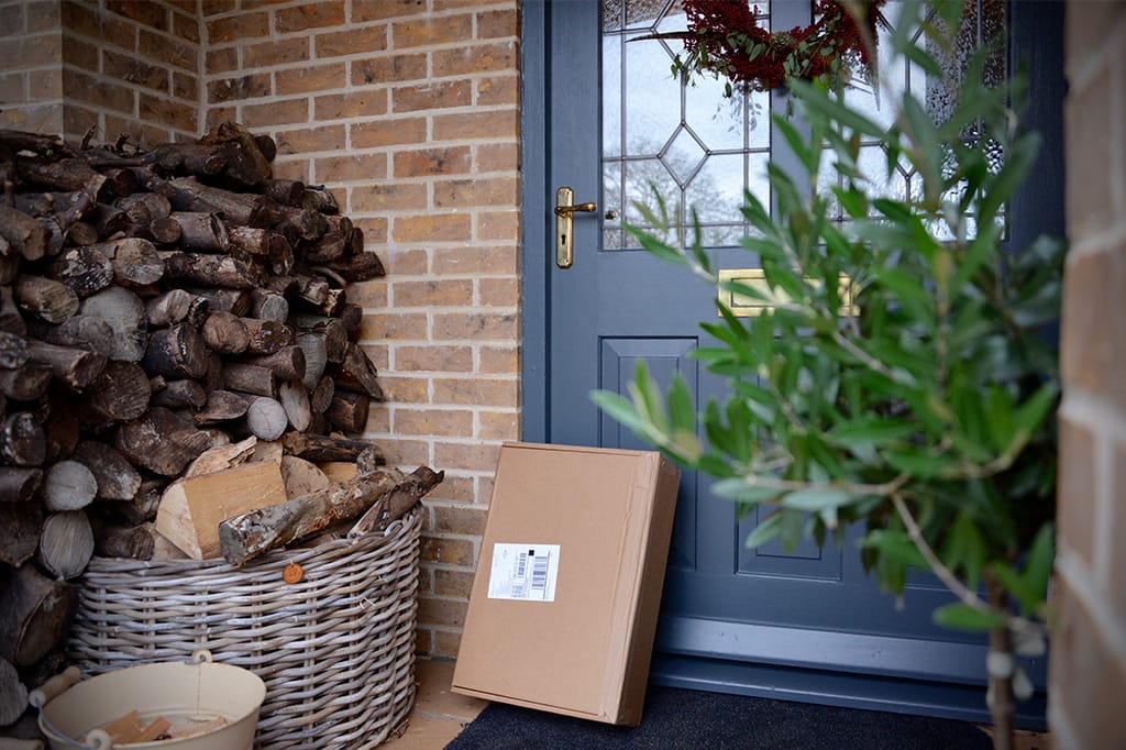 Items on doorstep left outside front door, next to firewood stack and plant – cardboard box delivery placed by blue door with wreath.